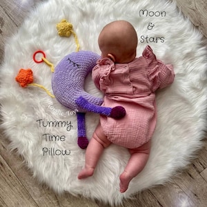 Könnte beinhalten: Ein Baby liegt auf einem flauschigen weißen Teppich neben einem lila gehäkelten mondförmigen Kissen mit geschlossenen Augen. Das Kissen ist mit "Tummy Time Pillow" beschriftet. Gelbe und orangefarbene gehäkelte Sterne sind befestigt. Der Text "Moon & Stars" ist ebenfalls sichtbar.