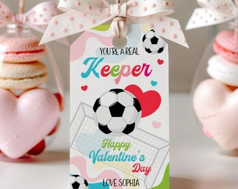 Puede incluir: Una etiqueta de regalo blanca con temática de fútbol, con un balón de fútbol, una portería y el texto "You're a Real Keeper" y "Happy Valentine's Day". La etiqueta está adornada con un lazo rosa con lunares.