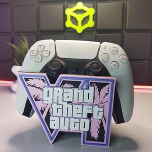 Puede incluir: Un mando de PlayStation 5 blanco con un soporte de logotipo de Grand Theft Auto V azul y rosa. El logotipo presenta palmeras y el texto "Grand Theft Auto" en blanco.