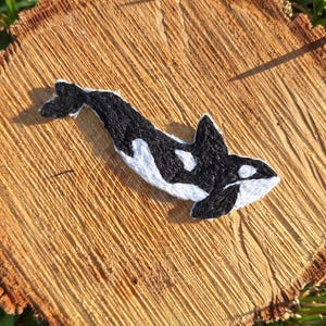 Pu&ograve; includere: Toppa ricamata di orca bianca e nera. L'orca sta nuotando verso destra. La toppa &egrave; su una superficie di legno.