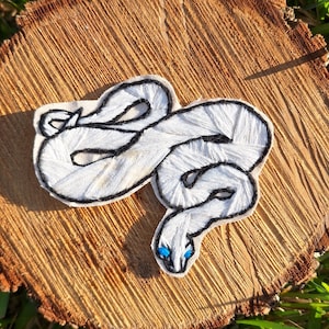 May include: White embroidered snake patch with blue eyes. The snake is curled up in a coil shape. The patch is made of white thread and has a black outline.