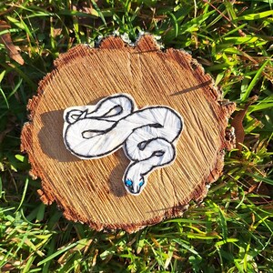 May include: White embroidered snake with blue eyes on a wooden background.