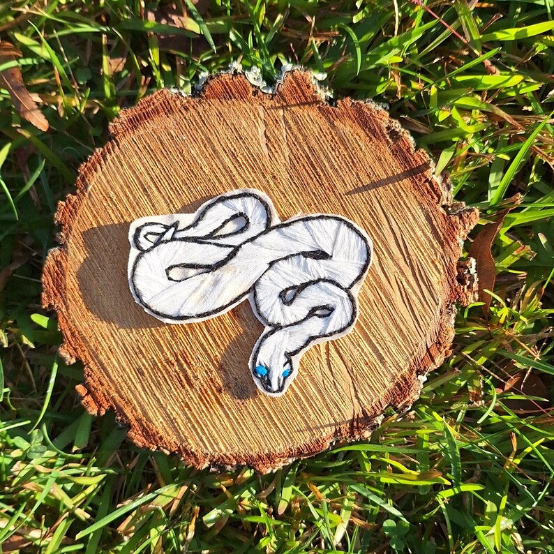 May include: White embroidered snake with blue eyes on a wooden background.
