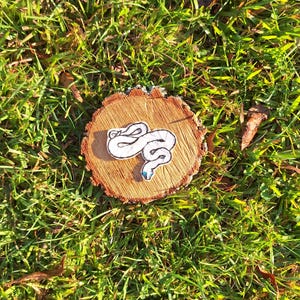 May include: White embroidered snake patch on a wooden tree stump. The snake is curled up with a blue eye.