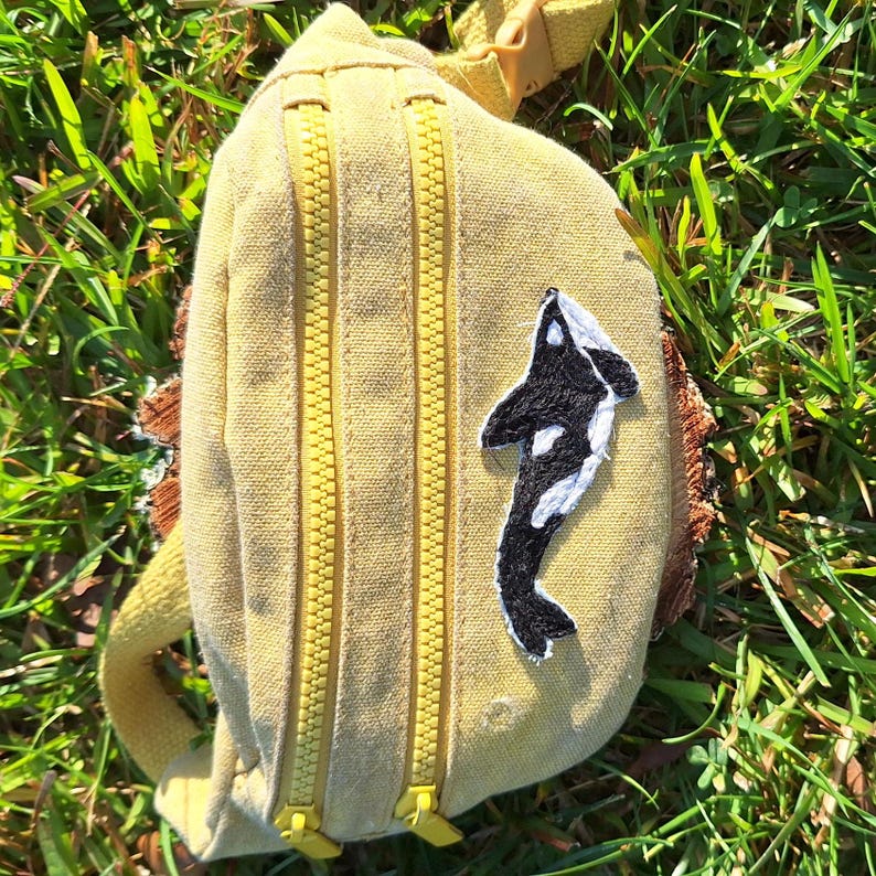 Pu&ograve; includere: Una marsupio in tela gialla con due cerniere gialle e una toppa di balena orca nera e bianca.