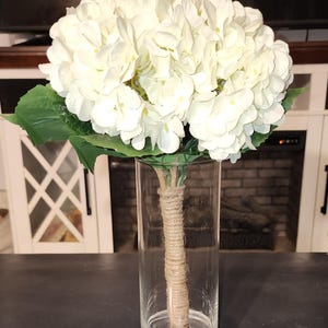 May include: A bouquet of white hydrangea flowers in a clear glass vase. The stems are wrapped in twine. The vase is cylindrical and sits on a dark surface. Green leaves are visible beneath the flower heads.