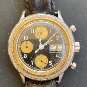 May include: A vintage Hamilton Chronograph watch with a black face and gold-toned subdials. The watch features a stainless steel case, a black leather strap, and the day and date displayed at the top. The watch hands are white.