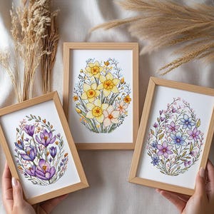 Könnte beinhalten: Drei gerahmte Blumen-Kreuzstich-Kunstwerke. Die Kunstwerke zeigen gelbe Narzissen, lila Krokusse und blaue und violette Blumen. Die Rahmen sind aus hellem Holz. Die Kunstwerke werden auf einem neutralen Hintergrund präsentiert.