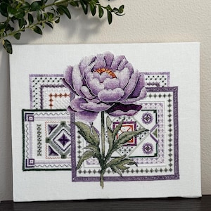 May include: A cross-stitch canvas featuring a large, purple peony flower with green leaves and stem. The flower is centered above three rectangular panels with intricate geometric patterns in shades of purple, green, and white. The canvas is mounted on a white background.