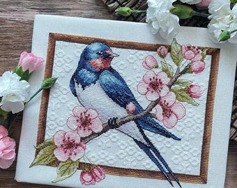 Op de afbeelding: Een borduurwerk met kruissteek van een blauwe en witte zwaluw die op een tak zit met roze bloesems. De zwaluw is gedetailleerd met een rode borst en een lange staart. De achtergrond is een witte stof met een subtiel patroon.