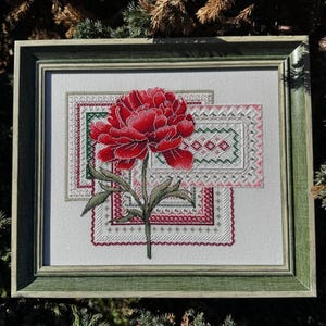 Puede incluir: Obra de arte en punto de cruz enmarcada con una peonía roja vibrante y hojas verdes. La flor está centrada sobre tres patrones rectangulares con detalles en rojo, verde y rosa. El marco es de color verde apagado.