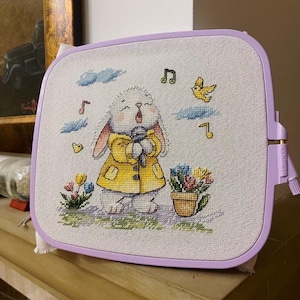 May include: A white bunny wearing a yellow jacket sings into a microphone with musical notes and a yellow bird flying above. The bunny is surrounded by flowers and a pot of flowers.