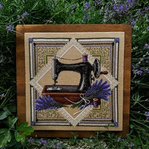 May include: A cross-stitch embroidery on beige fabric featuring a black vintage sewing machine, a bouquet of purple lavender, and a small spool of thread. The design is framed by a white lace-like border and intricate green and gold floral patterns.