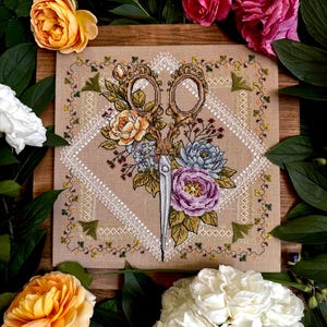 Könnte beinhalten: Ein quadratisches Stück beigefarbener Stoff mit einer detaillierten Kreuzstichstickerei, die eine verzierte Vintage-Schere zeigt, umrankt von bunten Blumen in Orange, Violett und Blau, eingefasst von einem weißen geometrischen Rautenmuster.