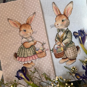 May include: Two embroidered cross-stitch panels featuring bunnies holding baskets of Easter eggs. The bunny on the left is wearing a green dress and the bunny on the right is wearing a green jacket and bow tie. Both panels have a light background with white polka dots.