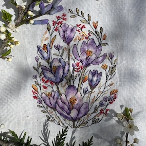 May include: A cross-stitch embroidery of a bouquet of purple crocuses with orange and red accents on a white linen background.