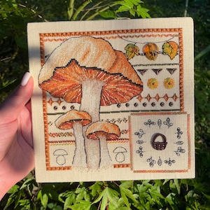 Puede incluir: Una bordado de punto de cruz con un grupo de setas de color naranja y blanco con una cesta marrón y hojas en el fondo. El bordado está enmarcado con un borde marrón.