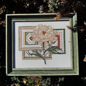 May include: A framed cross-stitch floral design featuring a large, cream-coloured peony with a yellow centre. The artwork is set against a white background, framed in a green wooden frame. The design includes geometric patterns in shades of green, red, and gold.