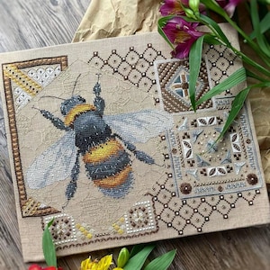 May include: A cross-stitch embroidery depicting a detailed bumblebee with yellow and black stripes, surrounded by geometric patterns in shades of brown, white, and grey.