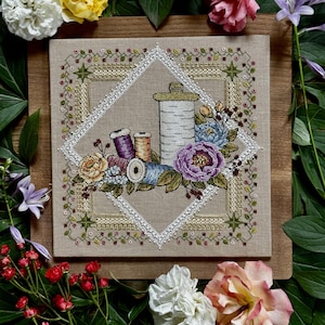 May include: A cross-stitch embroidery on beige fabric featuring a central diamond border enclosing spools of thread in purple, orange, and blue, surrounded by colorful floral arrangements of roses and peonies with green leaves and decorative stitching.
