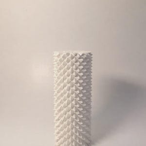 May include: A tall, cylindrical sculpture with a textured surface of repeating, white, triangular shapes. The geometric design creates a spiky, three-dimensional effect. The artwork stands on a white background.