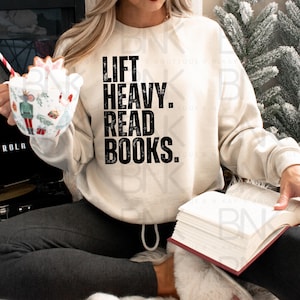May include: A woman wearing a cream colored sweatshirt with the text "Lift Heavy. Read Books." in black. She is holding a book in her lap.