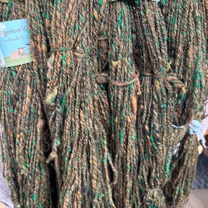 Puede incluir: Varias madejas de lana marrón gruesa y texturizada con motas verdes, amarillas y negras. Cada madeja está asegurada con una etiqueta de papel azul claro que muestra el texto Markree Wool Craft y una ilustración de una oveja en Sligo, Irlanda.