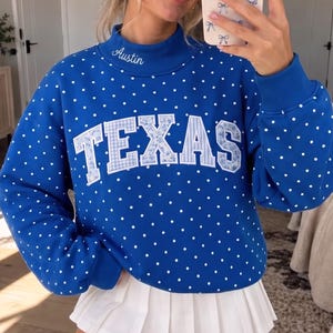 Könnte beinhalten: Ein königsblaues Sweatshirt mit Stehkragen, weißem Punktemuster und dem Wort TEXAS in großen Blockbuchstaben mit blau-weißem Blumen- und Vichy-Karo-Muster. Das Wort Austin ist in weißer Schrift auf den Stehkragen gestickt.