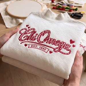 Könnte beinhalten: Ein weißes Sweatshirt mit Stehkragen und roter Vichy-Stickerei mit der Aufschrift Chi Omega und EST. 1895, akzentuiert durch kleine rote gestickte Herzen. Der Stoff ist eine weiche Baumwollmischung mit geripptem Kragen.