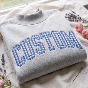 Op de afbeelding: Een lichtgrijs sweatshirt met ronde hals en het woord CUSTOM in grote blokletters. De letters zijn gevuld met een blauw-wit vichy-ruitpatroon en omlijnd met blauwe draad, wat zorgt voor een opvallende, gestructureerde uitstraling.
