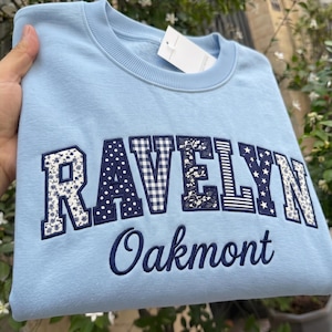 Könnte beinhalten: Ein hellblaues Sweatshirt mit Rundhalsausschnitt, das das Wort RAVELYN in großen Blockbuchstaben mit verschiedenen marineblauen und weißen Mustern wie Blumen, Karos, Punkten und Sternen zeigt, mit dem darunter in Marineblau gestickten Wort Oakmont.