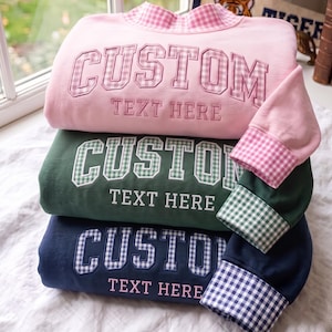Puede incluir: Una pila de tres sudaderas de manga larga en rosa claro, verde oscuro y azul marino. Cada sudadera presenta la palabra CUSTOM en una aplicación con patrón de cuadros vichy y el texto TEXT HERE bordado debajo. Los puños y cuellos tienen vichy a juego.