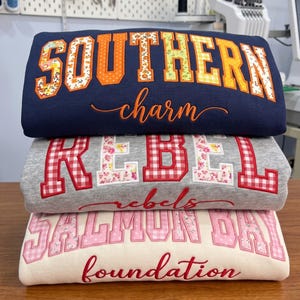 May include: A stack of three folded sweatshirts. The top navy blue sweatshirt features the text SOUTHERN in orange floral applique and charm in orange script. The middle gray sweatshirt has REBEL in red gingham and floral applique and rebels in red script. The bottom cream sweatshirt has SALMON BAY in pink gingham and floral applique and foundation in red script.