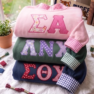 Könnte beinhalten: Ein Stapel aus drei langärmeligen Sweatshirts in Rosa, Dunkelgrün und Marineblau. Jedes Sweatshirt zeigt große griechische Buchstaben-Applikationen mit verschiedenen Blumen-, Punkt- und Streifenmustern. Kragen und Ärmelbündchen haben ein passendes Vichy-Karo-Muster.