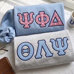 May include: A light blue sweatshirt featuring pink Greek letters with white polka dots and a dark blue border, paired with a heather gray sweatshirt displaying light blue Greek letters with white polka dots and a dark blue border.