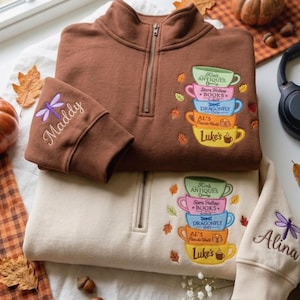 Puede incluir: Una sudadera marrón y otra crema con cremallera de cuarto, cada una con una pila bordada de cinco tazas coloridas etiquetadas como Kim's Antiques, Stars Hollow Books, Dragonfly Inn, Al's Pancake World y Luke's, con una libélula y un nombre en la manga.