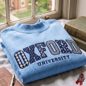 Könnte beinhalten: Ein hellblaues Sweatshirt mit Rundhalsausschnitt, das den Schriftzug OXFORD in großen, gemusterten Stoffbuchstaben mit verschiedenen Designs wie Vichy-Karo, Punkten und Sternen zeigt, darunter das Wort UNIVERSITY in marineblauem Garn.