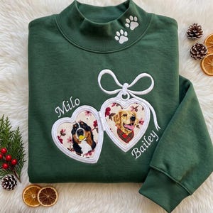 May include: A dark green mock neck sweatshirt featuring embroidered white paw prints on the collar and two heart-shaped locket designs on the chest. The left locket contains a portrait of a dog named Milo, and the right locket shows a dog named Bailey.