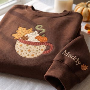 May include: A dark brown crewneck sweatshirt featuring an embroidered applique of a mug filled with whipped cream, a pumpkin, a star anise, and a maple leaf. The sleeve is embroidered with the name Maddy and a small leaf icon.