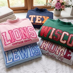 Puede incluir: Una pila de cinco sudaderas de cuello alto en rosa, azul marino, verde, azul claro y gris. Cada sudadera presenta grandes letras de bloque aplicadas en tela con varios patrones florales, de cuadros vichy y lunares, mostrando textos como LIONS, TEX, WISCON, LEWIS y HOME.