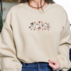 K&ouml;nnte beinhalten: Beiges Sweatshirt mit gesticktem Halloween-Design. Das Design zeigt vier Cartoon-Geister, einen K&uuml;rbis und Herbstbl&auml;tter. Ein Geist tr&auml;gt einen Hexenhut. Das Sweatshirt hat lange &Auml;rmel und einen Rundhalsausschnitt.