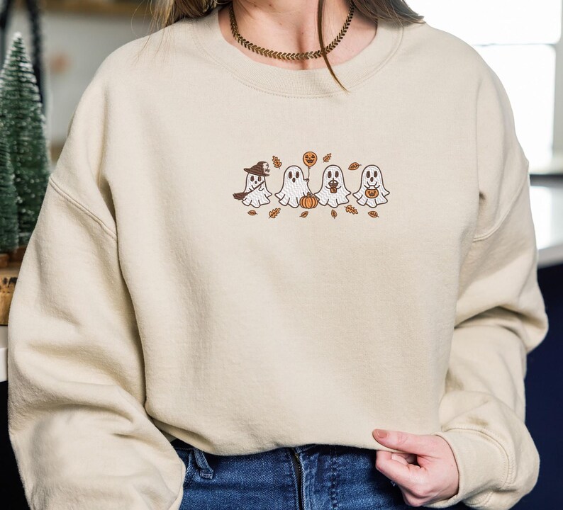 K&ouml;nnte beinhalten: Beiges Sweatshirt mit gesticktem Halloween-Design. Das Design zeigt vier Cartoon-Geister, einen K&uuml;rbis und Herbstbl&auml;tter. Ein Geist tr&auml;gt einen Hexenhut. Das Sweatshirt hat lange &Auml;rmel und einen Rundhalsausschnitt.