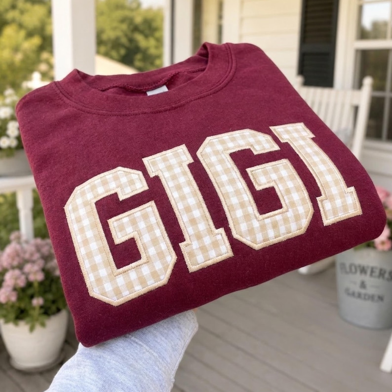 May include: A maroon crewneck sweatshirt featuring the word GIGI in large, block-style letters. The letters are filled with a beige and white gingham check pattern and are outlined with a thick, stitched border in a matching beige fabric.