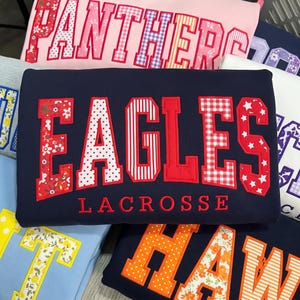 以下が含まれることがあります： ネイビーブルーのトレーナーで、赤と白の様々な柄の大きなブロック体で「EAGLES」と書かれ、その下に赤い刺繍で「LACROSSE」と記されています。文字には花柄、水玉模様、ストライプなどのパターンが施されています。