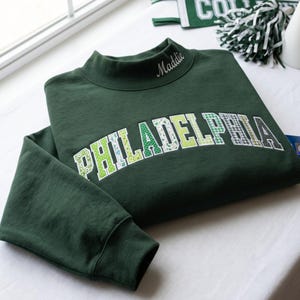 Puede incluir: Una sudadera verde oscuro de cuello alto con la palabra PHILADELPHIA en letras de bloque arqueadas y estampadas con diversos diseños en verde y blanco. El nombre Maddie está bordado en cursiva blanca en el lado derecho del cuello.