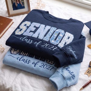 May include: A navy blue sweatshirt with the word SENIOR in patterned blue and white letters and the text class of 2027, plus a light blue sweatshirt with similar text. Both feature embroidered names on the sleeves, Stephanie and Charlotte.