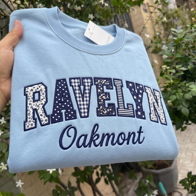 May include: A light blue crewneck sweatshirt featuring the word RAVELYN in large, block-style letters with varied navy blue and white patterns, including floral, gingham, polka dot, and star prints, with the word Oakmont embroidered below in navy.