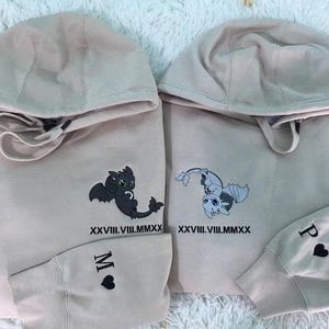 Może przedstawiać: Dwie jasnobeżowe bluzy z kapturem z czarno-białym nadrukiem smoka na każdej. Bluza z kapturem po lewej stronie ma czarnego smoka i napis "XXVIII.VIII.MMXX" oraz "M" na rękawie. Bluza z kapturem po prawej stronie ma białego smoka i napis "XXVIII.VIII.MMXX" oraz "P" na rękawie.