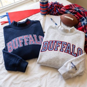 Puede incluir: Dos sudaderas de cuello alto, una azul marino y otra gris jaspeado, ambas con la palabra BUFFALO en letras de aplique de cuadros vichy rojos y azules. La sudadera azul marino tiene 716 bordado en el cuello y la gris western ny en la manga.