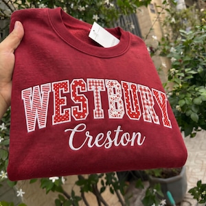 May include: A maroon crewneck sweatshirt featuring the word WESTBURY in large, block-style letters, each with a unique red and white pattern including stripes, polka dots, stars, and gingham. Below, the word Creston is embroidered in white script.